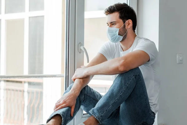 Homem com máscara médica sentado no peitoril da janela em casa — Fotografia de Stock
