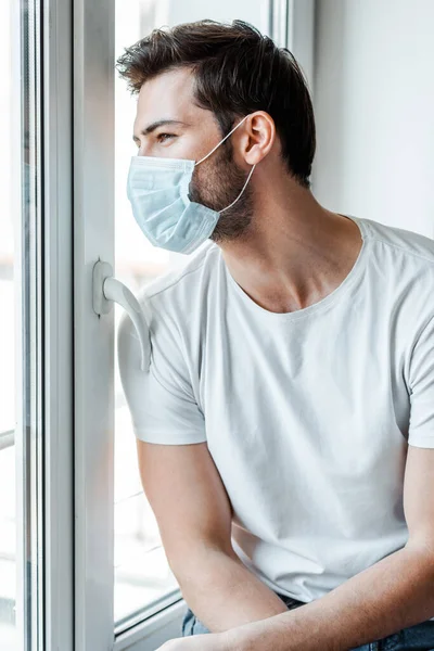 Mann mit medizinischer Maske schaut zu Hause auf Fenster — Stockfoto