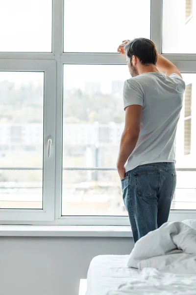 Селективный фокус человека с рукой в кармане, смотрящего в окно в спальне — стоковое фото