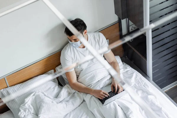 Vista aérea del hombre en máscara médica usando el ordenador portátil en la cama - foto de stock