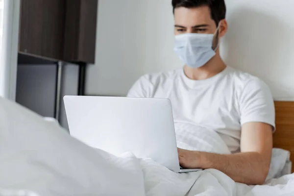 Selective focus of freelancer in medical mask working with laptop on bed — Stock Photo
