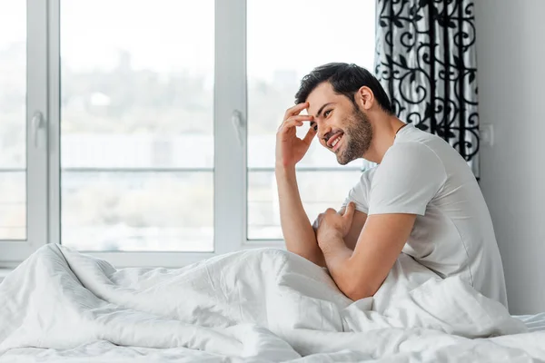 Seitenansicht eines gutaussehenden Mannes, der auf dem Bett sitzt und weglächelt — Stockfoto