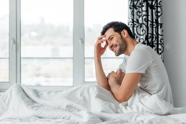 Vue latérale du bel homme souriant tout en étant assis sur le lit — Photo de stock