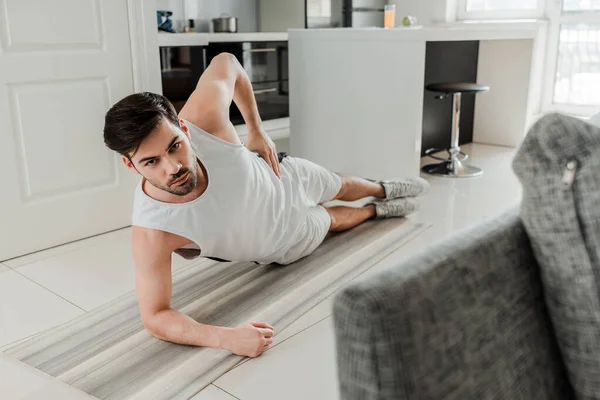 Selektiver Fokus des Mannes, der in die Kamera schaut, während er zu Hause auf der Fitnessmatte plankt — Stockfoto