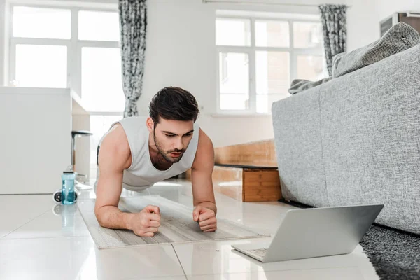 Vista de ángulo bajo del hombre mirando el ordenador portátil mientras hace tablón en la estera de fitness en casa - foto de stock