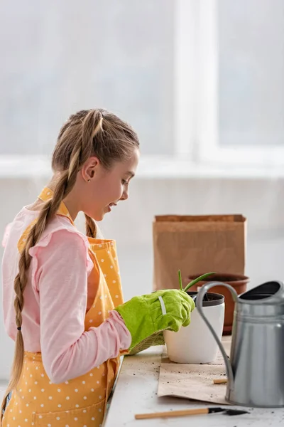 Seitenansicht des niedlichen Kindes mit Harke lächelnd und Blick auf Blumentopf mit Gießkanne am Tisch in der Küche — Stockfoto