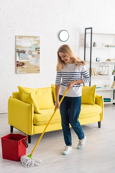 Hermosa mujer limpiando con fregona y sonriendo en la sala de estar - foto de stock