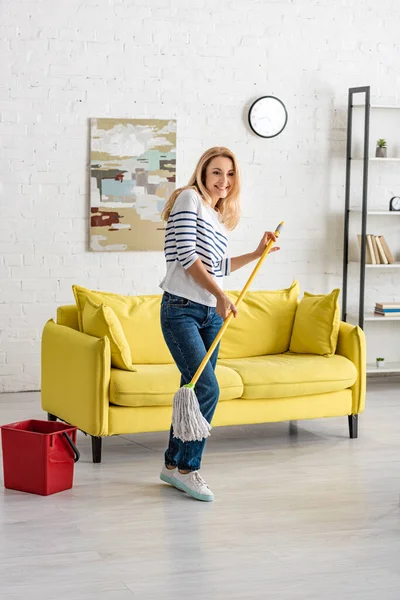 Beautiful woman holding mop near bucket and smiling in living room — Stock Photo