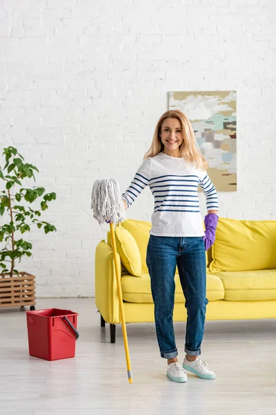 Beautiful woman with hand on hip and mop looking at camera and smiling near bucket in living room — Stock Photo