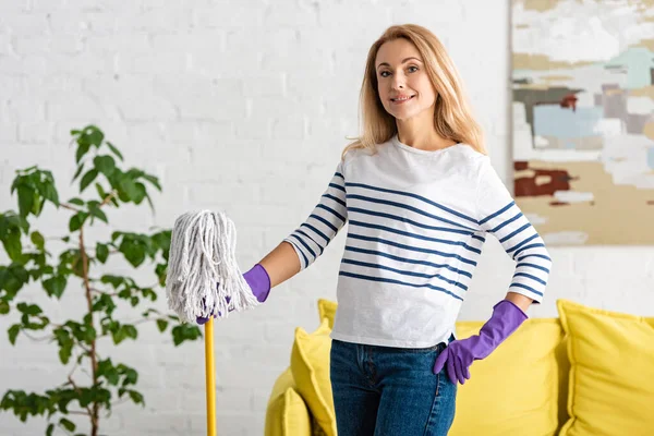 Woman in rubber gloves with hand on hip and mop looking at camera and smiling in living room — Stock Photo