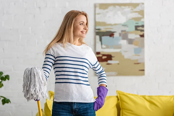 Frau in Gummihandschuhen mit Hand an Hüfte und Wischmopp, die im Wohnzimmer wegschaut und lächelt — Stockfoto