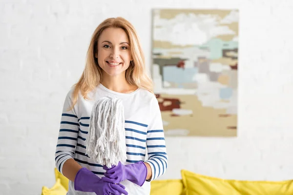 Frontansicht einer blonden Frau in Gummihandschuhen, die lächelt, in die Kamera blickt und im Wohnzimmer einen Wischmopp hält — Stockfoto