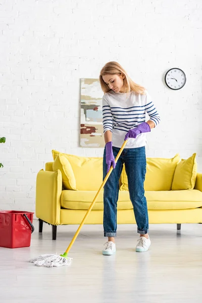 Woman in rubber gloves mopping floor near bucket in living room — Stock Photo