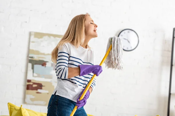 Woman in rubber gloves with closed eyes holding mop and singing in living room — Stock Photo