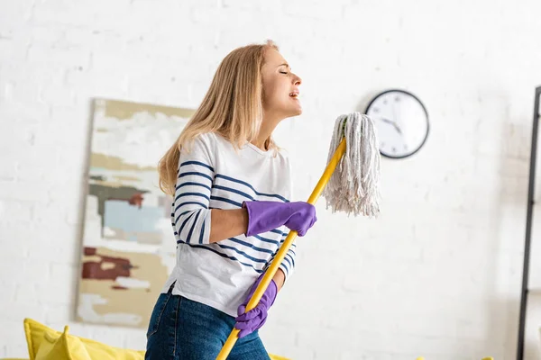 Frau in Gummihandschuhen mit geschlossenen Augen hält Wischmopp in der Hand und singt im Wohnzimmer — Stockfoto