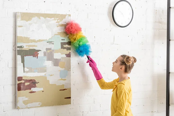Niedliches Kind in Gummihandschuhen fegt Staub vom Bild mit Staubwedel im Wohnzimmer — Stockfoto