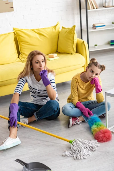 High angle view of tired mother and daughter with crossed legs holding mop with feather duster near dustpan on floor — Stock Photo