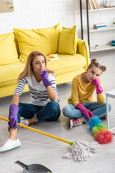 Vista de alto ângulo da mãe cansada e filha com pernas cruzadas segurando esfregona com espanador de penas perto de dustpan no chão — Fotografia de Stock