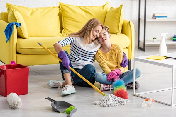 Müde Mutter umarmt Tochter mit Putzutensilien auf dem Boden neben Couchtisch im Wohnzimmer — Stockfoto