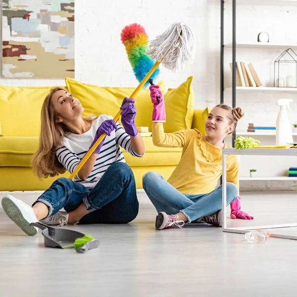 Madre e hija peleando con trapeador y plumero y sonriendo cerca de la mesa de café y cepillo con recogedor en el suelo en la sala de estar - foto de stock