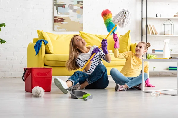 Madre e hija peleando con fregona y plumero cerca de mesa de café y artículos de limpieza en el suelo en la sala de estar — Stock Photo