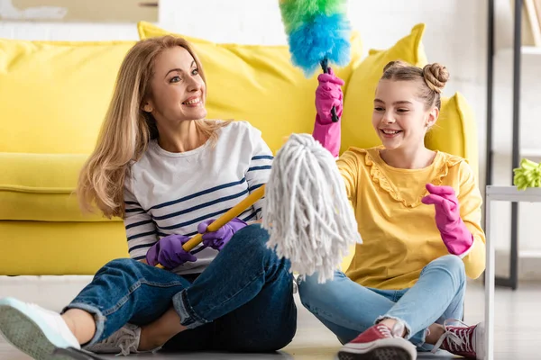 Mother and cute daughter with mop and feather duster smiling on floor in living room — Stock Photo