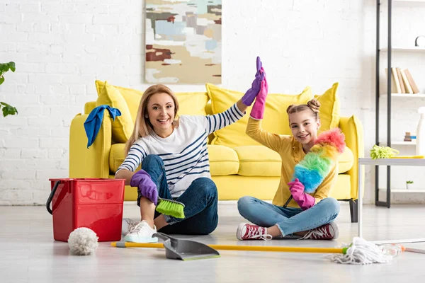 Madre y linda hija con suministros de limpieza dando choco cinco, sonriendo y mirando a la cámara en el suelo en la sala de estar - foto de stock