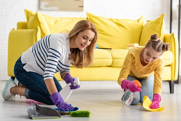 Madre y linda hija con botellas de spray y trapos que limpian el suelo cerca del cepillo y el recogedor en la sala de estar - foto de stock