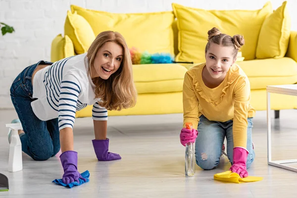 Madre a quattro zampe e figlia con bottiglie spray e stracci che puliscono il pavimento, sorridendo e guardando la fotocamera in soggiorno — Foto stock