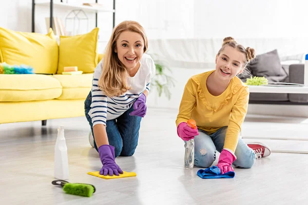 Madre e hija con botella de spray y trapos limpiando el suelo, sonriendo y mirando a la cámara cerca del cepillo en la sala de estar — Stock Photo