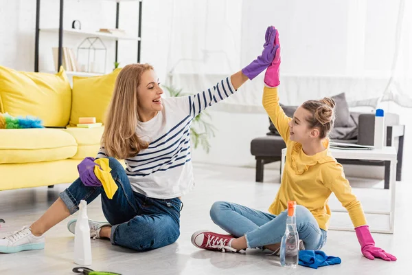 Madre e figlia con prodotti per la pulizia dando il cinque, sorridendo e guardandosi l'un l'altro sul pavimento in soggiorno — Foto stock