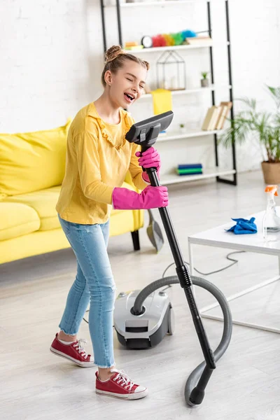 Cute child with closed eyes singing with vacuum cleaner near coffee table in living room — Stock Photo