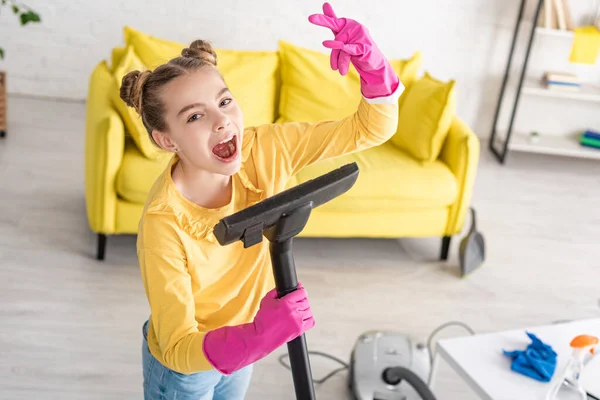 Vue en angle élevé de l'enfant mignon chantant avec aspirateur et montrant signe de roche dans le salon — Photo de stock