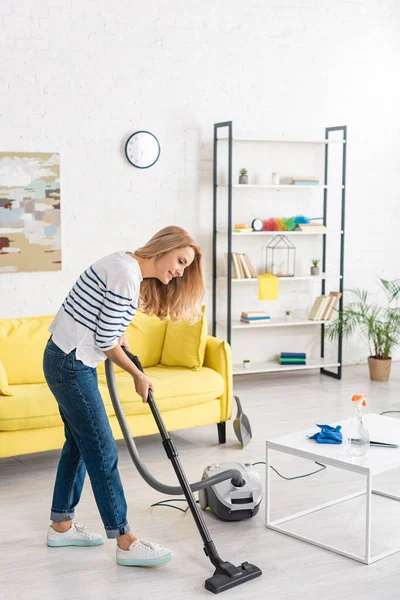 Frau räumt mit Staubsauger neben Couchtisch und Sofa im Wohnzimmer auf — Stockfoto