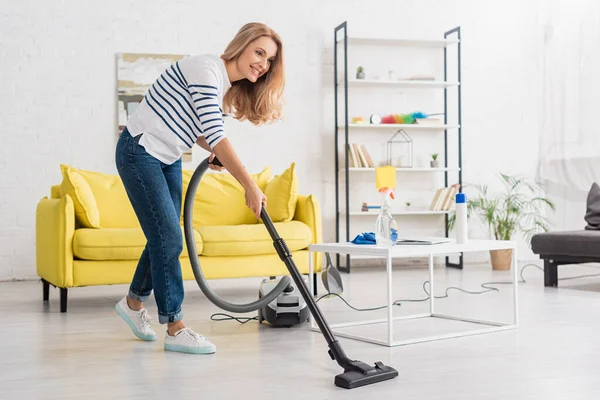 Woman smiling and cleaning up with vacuum cleaner near coffee table and sofa in living room — Stock Photo