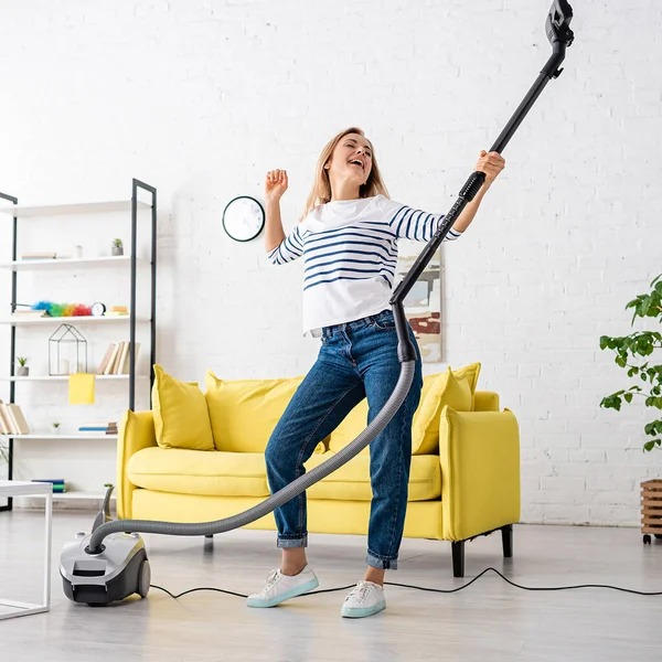 Blonde woman singing with closed eyes and vacuum cleaner near sofa in living room — Stock Photo