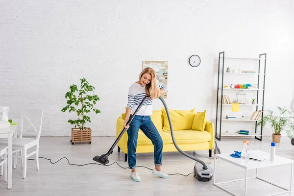 Mujer rubia sonriendo y limpiando con aspiradora en el salón - foto de stock