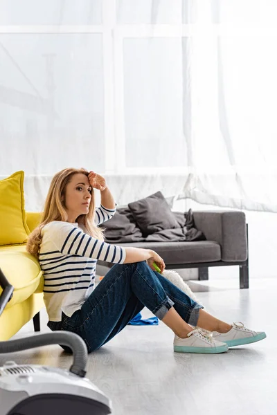 Selektiver Fokus einer müden Frau mit Staubwedel, die neben Sofa und Staubsauger auf dem Boden im Wohnzimmer sitzt — Stockfoto