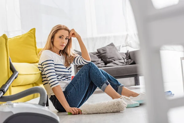 Selective focus of tired woman with feather duster looking at camera and sitting near sofa and vacuum cleaner on floor in living room — Stock Photo