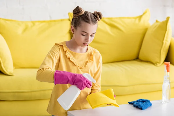 Cute child with spray bottle applying detergent on rag near coffee table and sofa in living room — Stock Photo