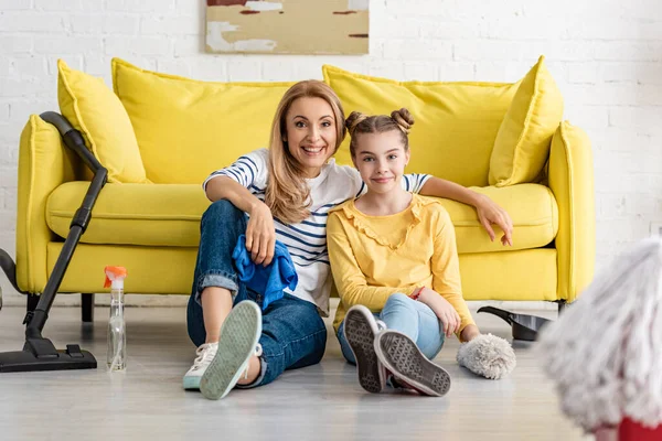Focus selettivo di madre e figlia carina guardando la fotocamera e sorridendo vicino al divano e forniture per la pulizia sul pavimento in soggiorno — Foto stock