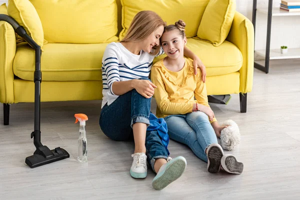 Madre abrazando linda hija y sonriendo cerca de sofá y artículos de limpieza en el suelo en la sala de estar - foto de stock