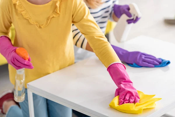 Vista recortada de la madre y la hija con botellas de spray limpiando la mesa de café con trapos en la sala de estar - foto de stock