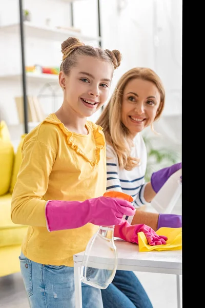 Cute child holding rag and spray bottle, smiling and looking at camera with mother near coffee table in living room — Stock Photo