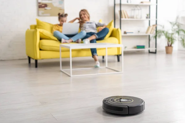 Selective focus of mother and daughter with laptop on sofa near coffee table and robotic vacuum cleaner on floor in living room — Stock Photo