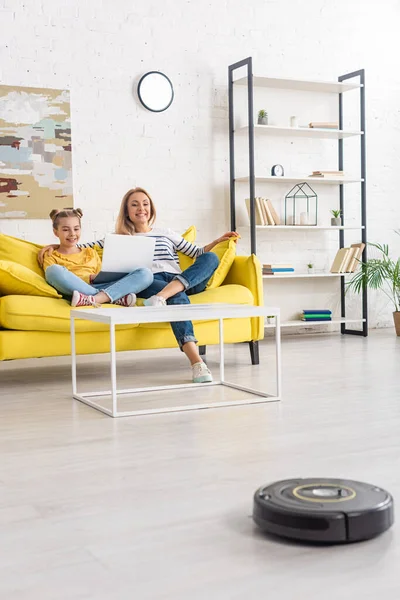 Mother smiling, hugging daughter with laptop on sofa near coffee table and looking at robotic vacuum cleaner on floor in living room — Stock Photo