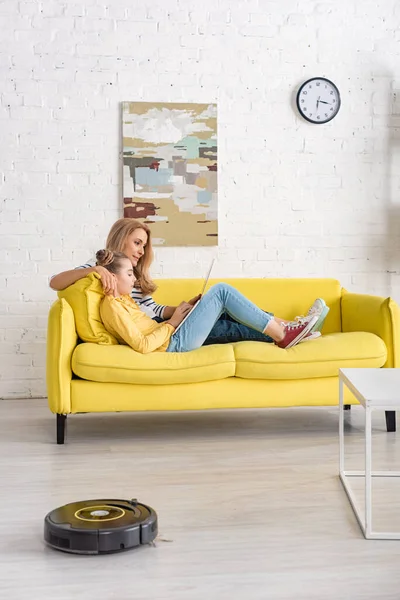 Mère et fille avec ordinateur portable couché sur le canapé près de la table basse et aspirateur robotisé sur le sol dans le salon — Photo de stock