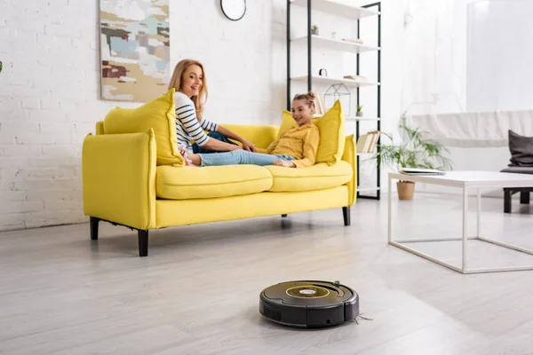 Mère et fille souriantes et allongées sur le canapé près de la table basse et aspirateur robotisé sur le sol dans le salon — Photo de stock