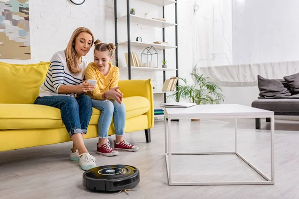 Mãe e filha olhando para o smartphone no sofá perto da mesa de café com laptop e aspirador robótico no chão na sala de estar — Fotografia de Stock