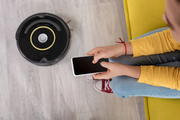 Обрезанный вид ребенка со смартфоном на диване рядом с роботизированным пылесосом на полу в гостиной — стоковое фото